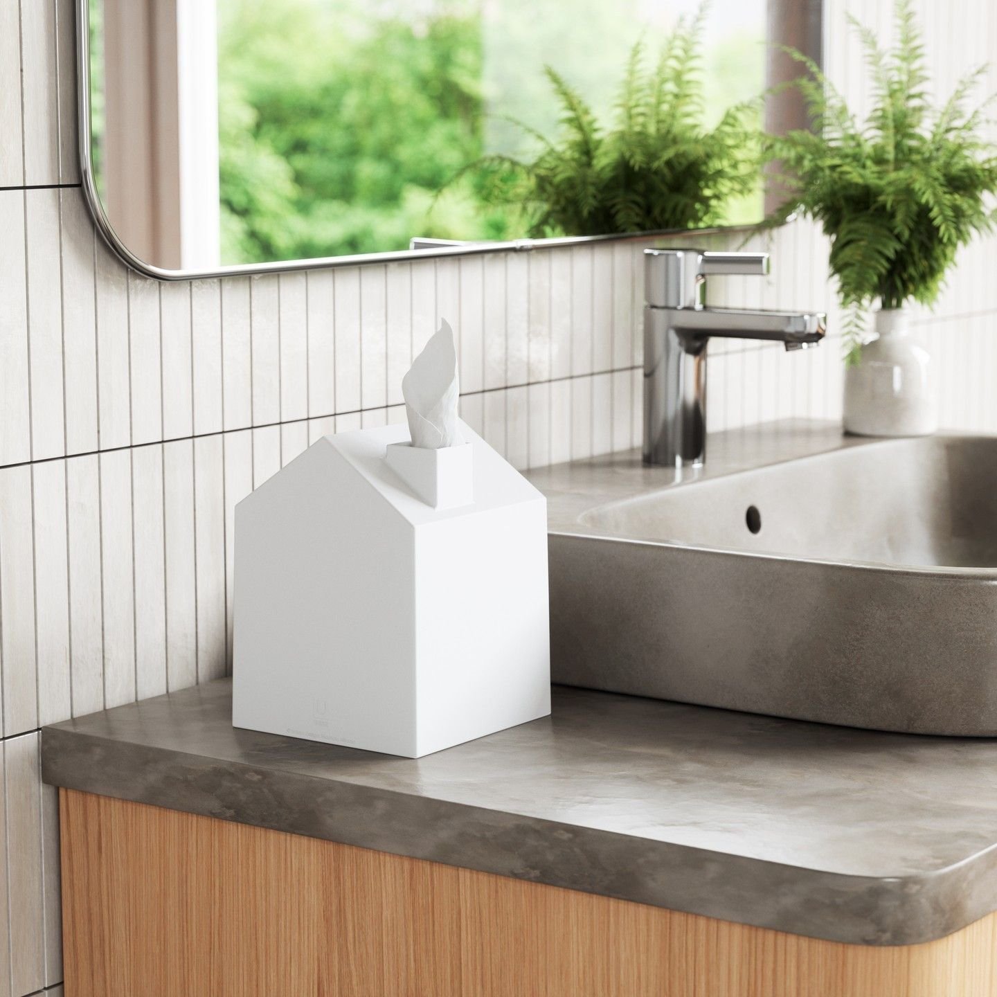 Umbra  Casa Tissue Cover on a bathroom counter with a sink next to it. A tissue is sticking out the top of the box